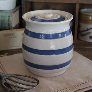 May include: A white ceramic jar with blue horizontal stripes and a matching lid. The jar is cylindrical with a rounded base and is sitting on a beige cloth. A vintage whisk is in the foreground.