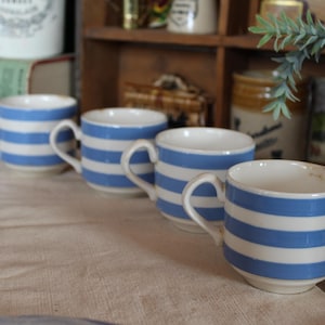 Può includere: Quattro tazze in ceramica a righe orizzontali blu e bianche sono disposte su un runner in lino beige. Le tazze hanno manici bianchi. Lo sfondo presenta oggetti vintage, suggerendo un'estetica rustica.