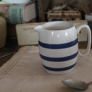 Peut inclure: Une cruche en céramique blanche avec trois rayures bleues horizontales. La cruche a une anse et un bec verseur. Elle est posée sur un tissu beige à bord décoratif, à côté d'une cuillère.