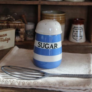 May include: A vintage sugar shaker with blue and white stripes and the word "SUGAR" in black. It sits on a white cloth with a metal whisk. A box of "Clotted Cream Fudge" is in the background.