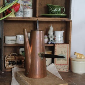 May include: A copper-coloured vintage coffee pot with a black handle and spout. The pot is tall and slender, with a pointed top. It sits on a stack of books, with a wooden shelf in the background filled with various antique items.