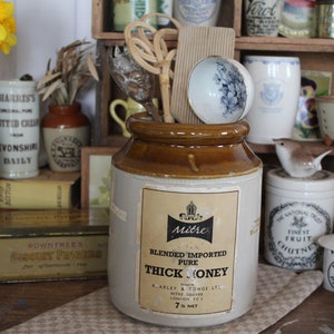 Może przedstawiać: Cylindryczny kamionkowy garnek z brązowym glazurowanym brzegiem i kremowym korpusem. Papierowa etykieta zawiera napis Mitre Blended Imported Pure Thick Honey, pakowany przez K. Arley & Tonge Ltd, Mitre Square, London EC3, 7 lb net. Wewnątrz znajdują się przybory kuchenne.
