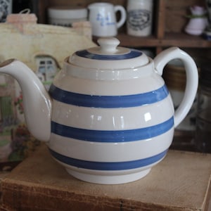 May include: A white ceramic teapot with horizontal blue stripes. The teapot has a curved spout, a rounded body, and a handle. It sits on a stack of books. The background includes jars and other kitchenware.