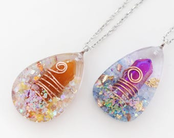Op de afbeelding: Twee druppelvormige hangerkettingen. Eén hanger heeft een oranje kristal gewikkeld in koperdraad, de andere een paars kristal. Beide zijn ingekapseld in heldere hars met glitter en hangen aan zilveren kettingen.