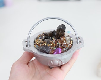May include: A small, silver-colored cauldron filled with a variety of crystals and gemstones. The crystals are dark brown, clear, and purple, and the gemstones are iridescent. The cauldron has a silver handle.