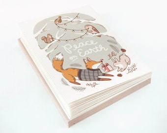 May include: A stack of greeting cards with a whimsical illustration. The design features a fox in a sweater, a rabbit, and other woodland creatures. The words "Peace on Earth" are written in a script font. The cards have a light gray background.