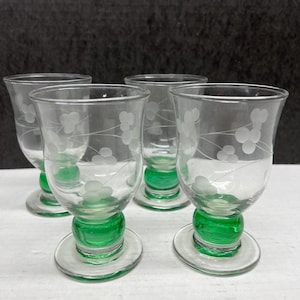 May include: Four clear glass cordial glasses with a green base and etched floral design. The glasses have a rounded base and a flared top. The green base sits on a clear, round foot.