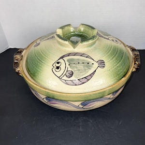 May include: Ceramic serving dish with a lid, featuring a hand-painted fish design. The dish has a green and cream color scheme with brown handles. The lid has a built-in ashtray.
