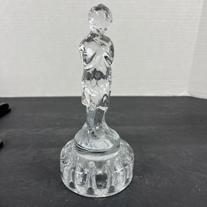May include: A clear glass figurine of a person with arms crossed, standing atop a round, textured base. The figure is detailed with visible facial features and clothing. The base has a series of vertical indentations around its perimeter.