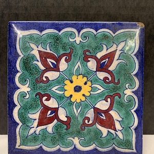 May include: Square ceramic tile with a floral design. The tile features a blue border, a green background, and a central floral pattern with red, white, and yellow accents. The design is symmetrical and detailed, suitable for decorative use.