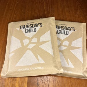 May include: Two beige paper packages with the text "THURSDAY'S CHILD" in black and white. The packages have a geometric design and the text "+OMORROW X +TOGETHER". The packages are on a wooden surface.