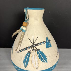 May include: A cream-colored ceramic vase with a narrow neck and a wide base. The vase features hand-painted blue and black geometric patterns, including feathers and a stylized axe. A blue leather cord with wooden and blue beads hangs from the neck.