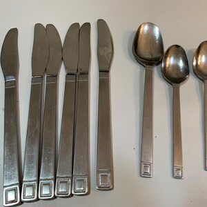 May include: A set of stainless steel flatware including butter knives and spoons. The knives have a rectangular design at the base of the handle. The spoons have a rounded bowl and a similar rectangular design.