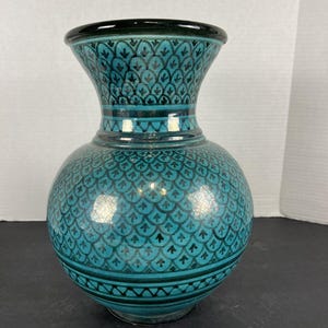 May include: A turquoise ceramic vase with a bulbous body and flared neck. The vase features a repeating pattern of stylized leaves in black and turquoise. The rim and base are dark green, and the vase has a glossy finish.