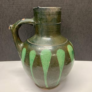 May include: A dark green ceramic pitcher with a handle and a spout. The pitcher is decorated with vertical green teardrop shapes. The handle has a small chip. The pitcher has a wide base and a narrow neck.