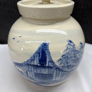 May include: A ceramic jar with a lid, featuring a hand-painted landscape scene in shades of blue and white. The jar has a round body and a lid with a small knob. The landscape depicts a building and trees.