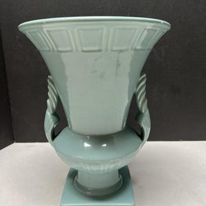 May include: A light blue ceramic vase with a square base and a bulbous midsection. The top flares out with a geometric pattern, and it has two decorative handles. The vase is approximately 10 inches tall.