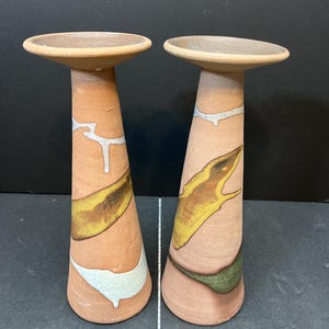May include: Two terracotta-colored candle holders with a wide, flat top and a flared base. Each holder features abstract designs in yellow, green, and white. The candle holders are displayed against a dark background.