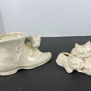 May include: Off-white ceramic planters. One planter is shaped like a boot with a dog figurine on top. The other planter features three cat figurines. These decorative planters are suitable for small plants or succulents.