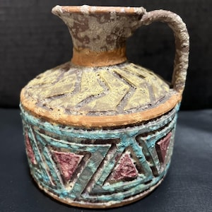 May include: A small, handcrafted ceramic pitcher with a handle. The pitcher features a textured surface with a geometric design in shades of yellow, teal, and red. The rim and handle are a darker brown.