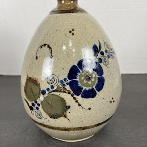 May include: A speckled, egg-shaped ceramic vase with a dark brown rim and neck. The vase features a blue floral design with green leaves and accents. The base has a dark brown band. The vase is a decorative item.