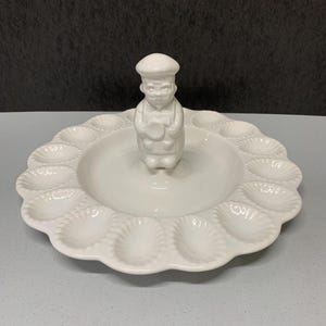 May include: White ceramic deviled egg serving platter with a scalloped edge and a small chef figurine in the center. The platter has 12 egg indentations around the edge.