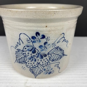 May include: A light gray stoneware crock featuring a hand-painted blue floral and strawberry motif on the front. The cylindrical vessel has a wide, flared rim and a smooth, speckled finish, showcasing a classic design with decorative blue accents.