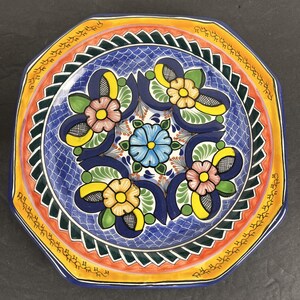 May include: An octagonal ceramic plate with a vibrant floral design. The plate features a blue center with a repeating pattern of flowers in pink, yellow, and blue, surrounded by orange, yellow, and green borders. The plate has a glossy finish.
