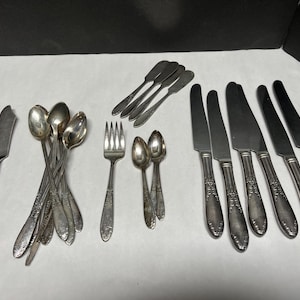 May include: A collection of antique silverware, including spoons, forks, knives, and butter knives. The silverware is silver-colored and arranged on a white surface. The utensils have decorative handles.