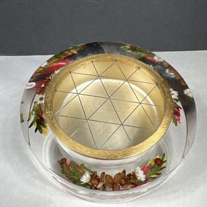 May include: A clear, round floral frog with a gold-colored metal grid and a gold-colored interior. The clear base is embedded with dried flowers and pinecones. A decorative piece for floral arrangements.