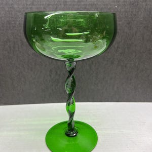 May include: A green glass goblet with a round bowl and a twisted stem. The base is a flat circle. The glass has a vibrant green color and is likely used for serving beverages.