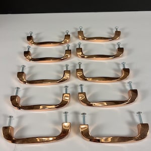 May include: Ten copper-colored cabinet pulls with a curved, U-shaped design. Each pull has two screw posts for easy installation. The pulls are arranged in two rows of five, showcasing their polished finish and hardware.