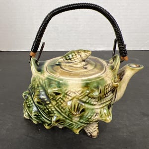 May include: A ceramic teapot with a seashell design in shades of green, tan, and brown. The teapot has a black woven handle and a spout. The lid is topped with a seashell. The teapot is approximately 6 inches tall.