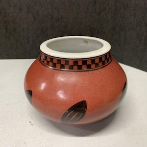 May include: A small, round ceramic pot with a speckled reddish-orange finish. The rim is white, and a band of black and brown checkered squares encircles the top. Black leaf-like shapes are painted on the sides.