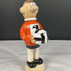 May include: A ceramic figurine of a soccer player in an orange jersey with the number 4, holding a black and white soccer ball. The figurine has a smiling face, a white collar, black shoes, and a beige base.