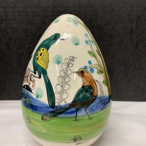 May include: A hand-painted ceramic egg with a whimsical design. The egg features two birds, one teal and yellow, the other brown and teal, set against a backdrop of blue, green, and white. Floral and foliage details complete the decorative piece.