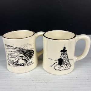 May include: Two cream-colored ceramic mugs with brown rim accents. The left mug features a black line drawing of the Berkeley Pit with the text 'BERKELEY PIT' and 'BUTTE, MONT'. The right mug shows a black illustration of a mine headframe with 'Butte, Montana'.