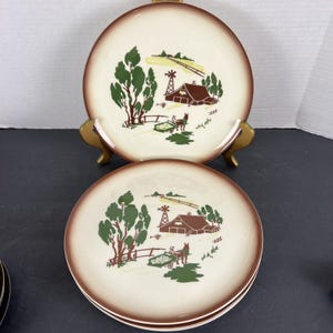May include: Set of vintage ceramic plates with a cream-colored center and a brown rim. Each plate features a rural scene with a barn, windmill, trees, and a horse-drawn cart. The plates are displayed on a gold-colored stand.