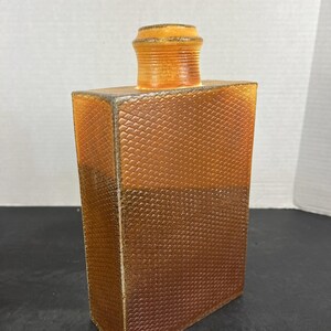 May include: A rectangular amber-colored glass bottle with a textured surface. The bottle has a short, cylindrical neck and a matching stopper. The bottle's design features a repeating pattern, and the edges are outlined with a silver trim.