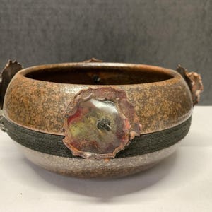 May include: A handcrafted ceramic bowl with a speckled brown exterior and a gray base. The bowl features decorative copper-colored accents and a black band around the middle. The interior is a warm orange color.