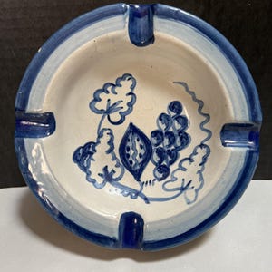May include: Round ceramic ashtray with a white interior and a blue and white painted design. The ashtray has a blue rim and four cigarette rests. The interior features a blue floral and grape design.