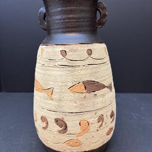 May include: A handcrafted ceramic vase with a dark brown top and handles. The beige body features brown fish and decorative patterns. The vase is approximately 10 inches tall, with a textured surface and a dark brown base.