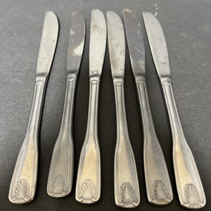 May include: Six stainless steel butter knives with serrated edges and decorative shell-shaped designs on the handles. The knives are arranged on a dark surface, showcasing their classic design and potential for elegant table settings.