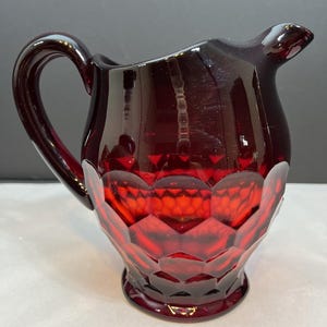 May include: A ruby red glass pitcher with a handle and spout. The pitcher has a faceted design on the lower half and a smooth, glossy finish. The pitcher is likely used for serving beverages.