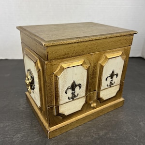 May include: A decorative wooden box with a gold finish and cream-colored panels. The box features a lion's head on one side and fleur-de-lis emblems. The box has a small drawer and a hinged lid.