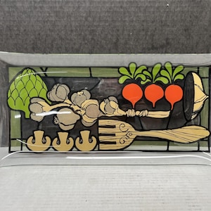 May include: A rectangular clear glass tray with a painted design of vegetables. The design includes an artichoke, garlic, carrots, mushrooms, and a wooden fork. The vegetables are in shades of green, orange, and tan.