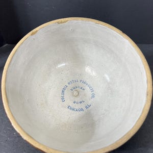 May include: A vintage ceramic bowl with a light beige interior and a brown rim. The bowl has the text "COLUMBIA METAL PRODUCTS CO. DUNLAP BOWL CHICAGO, ILL" printed in blue on the inside.