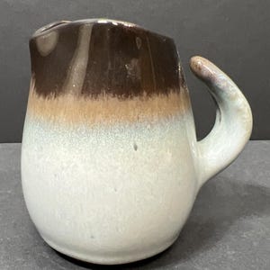 May include: A small, ceramic pitcher with a curved handle. The top is dark brown, transitioning to tan, then light blue, and finally white at the base. The handle is a light blue with brown accents.