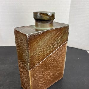 May include: A rectangular ceramic vase with a textured, scale-like pattern in shades of brown and gold. The top portion is a darker brown with a cylindrical opening. The vase has a diagonal color separation.
