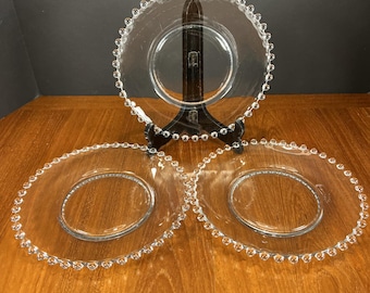 May include: Three clear glass plates with a beaded edge. One plate is displayed on a black stand, while the other two are lying flat on a wooden surface. The plates have a circular design with a raised center.